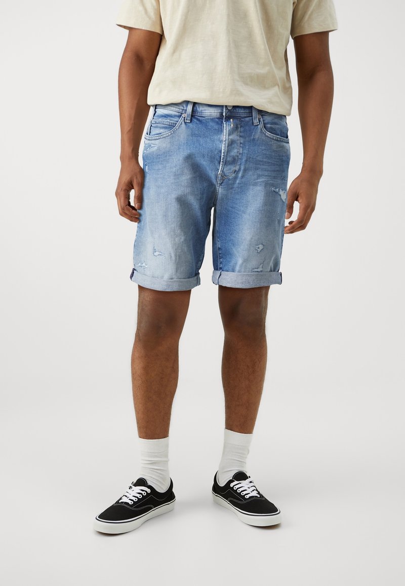 Homme portant un short en jean bleu clair retroussé, un t-shirt beige, des chaussettes blanches et des baskets noires basses, debout sur un fond uni.