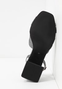 Sandale à talon haut grise avec un dessus en tissu texturé, bout carré et une semelle en caoutchouc noir robuste, dotée d'une sangle autour de la cheville et d'accents métalliques.