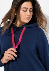 Felpa con zip di colore blu navy, dotata di un cappuccio tessurizzato, con lacci rosa, in morbido tessuto e con una vestibilità rilassata. Mano posizionata vicino alla zip.