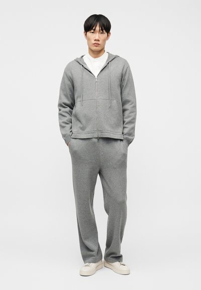 Νέος άνδρας που στέκεται με τα χέρια στις τσέπες, φορώντας γκρι φούτερ με φερμουάρ, γκρι παντελόνι που ταιριάζει και λευκά αθλητικά παπούτσια, σε λευκό φόντο.