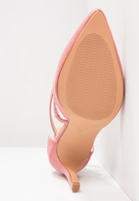 Chaussure à talon haut en daim rose avec bout pointu, bride à la cheville et semelle légèrement texturée, photographiée de dessous sur fond blanc.