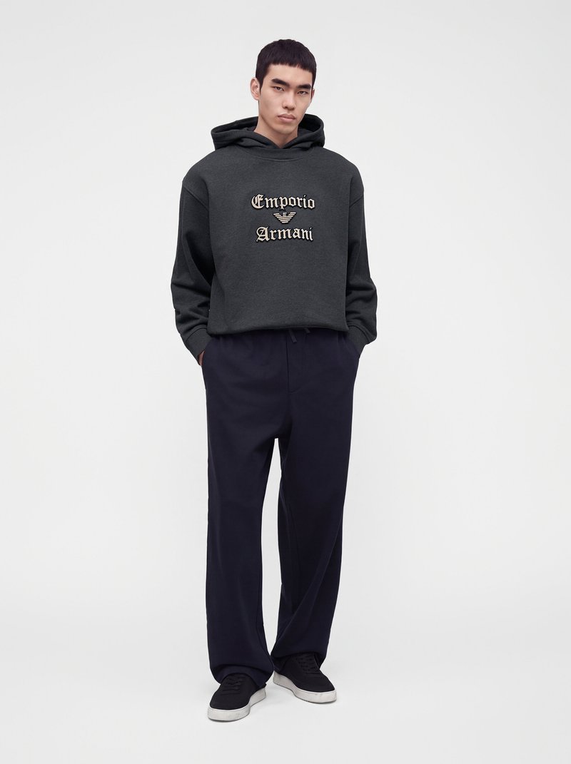 Млад мъж, облечен с тъмносива качулка Emporio Armani, широки черни панталони и черни кецове, стои на изчистен светъл фон.
