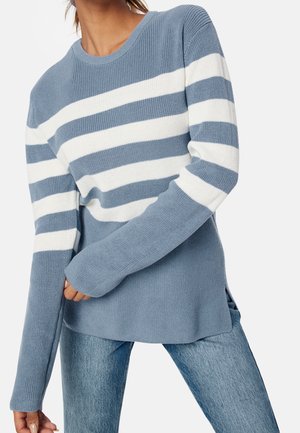 Femme vêtue d'un pull en maille rayé bleu et blanc à manches longues et d'un jean bleu, sur un fond uni.