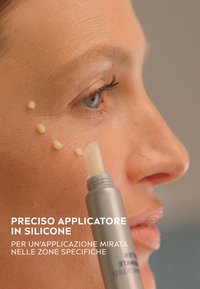 Applicatore in silicone con punta appuntita, erogando un prodotto beige chiaro sulla pelle. Texture liscia, design della confezione in argento e bianco.