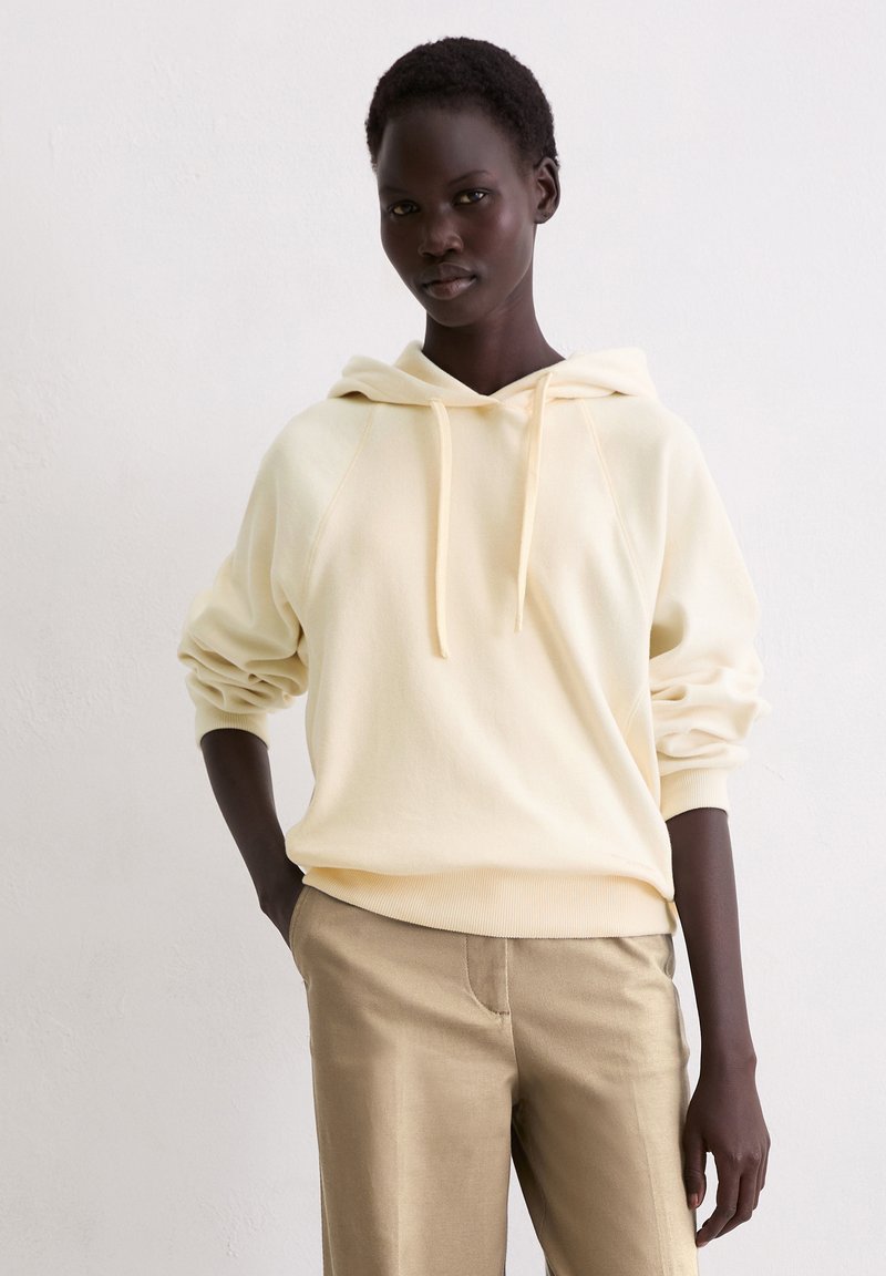 Crème hoodie met een relaxte pasvorm en ribgebreide boorden, gecombineerd met beige broek. Zachte textuur met trekkoorden bij de kap.