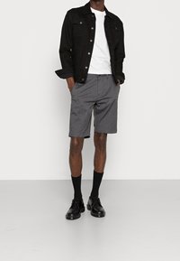 Homme portant une veste en denim noire, un t-shirt blanc, un short gris, des chaussettes noires et des chaussures en cuir noires, se tenant avec les mains dans les poches.