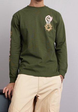 Homme portant une chemise à manches longues vert olive avec des motifs de paix et spirales roses et beiges, associée à un pantalon beige clair.