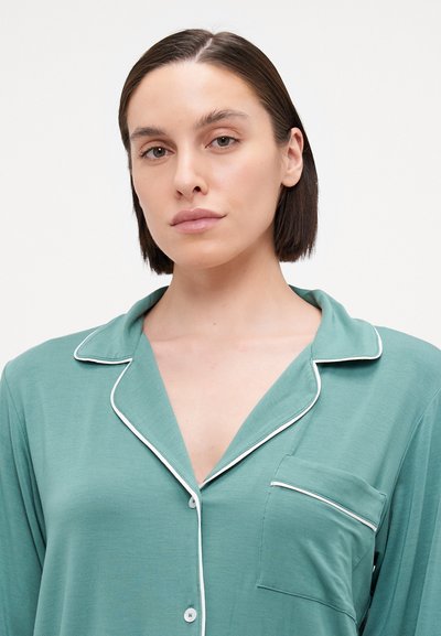 Light green pajama top with a collar, white piping, and a chest pocket. Fabric appears soft, with a smooth texture and relaxed fit.