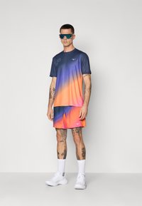 T-Shirt mit kurzen Ärmeln im Farbverlauf von Blau nach Orange, kombiniert mit passenden Shorts. Zubehör umfasst blaue Sonnenbrillen und weiße Sportschuhe.