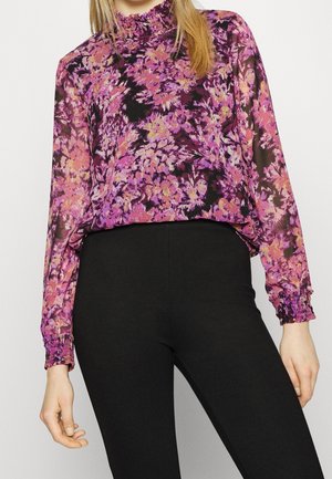 Blusa nera trasparente con motivo floreale viola, caratterizzata da un colletto alto arruffato e polsini elastici, abbinata a pantaloni neri aderenti.