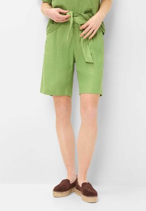 Personne portant un short en tissu vert clair avec une ceinture nouée à la taille, un haut vert assorti et des espadrilles marron à enfiler, debout devant un fond blanc.