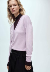 Ljuslila kofta med djup V-ringning, med långa ärmar och mörkbruna detaljer vid ärmsluten, matchad med en plisserad svart kjol.