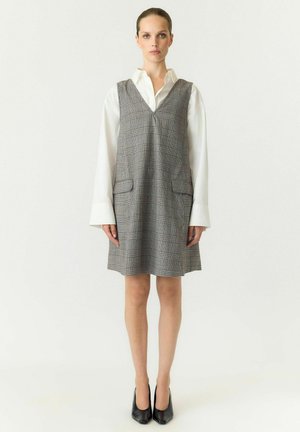 Femme portant une robe sans manches à carreaux gris par-dessus une chemise blanche à manches longues et des chaussures à talons noires, debout devant un fond uni.