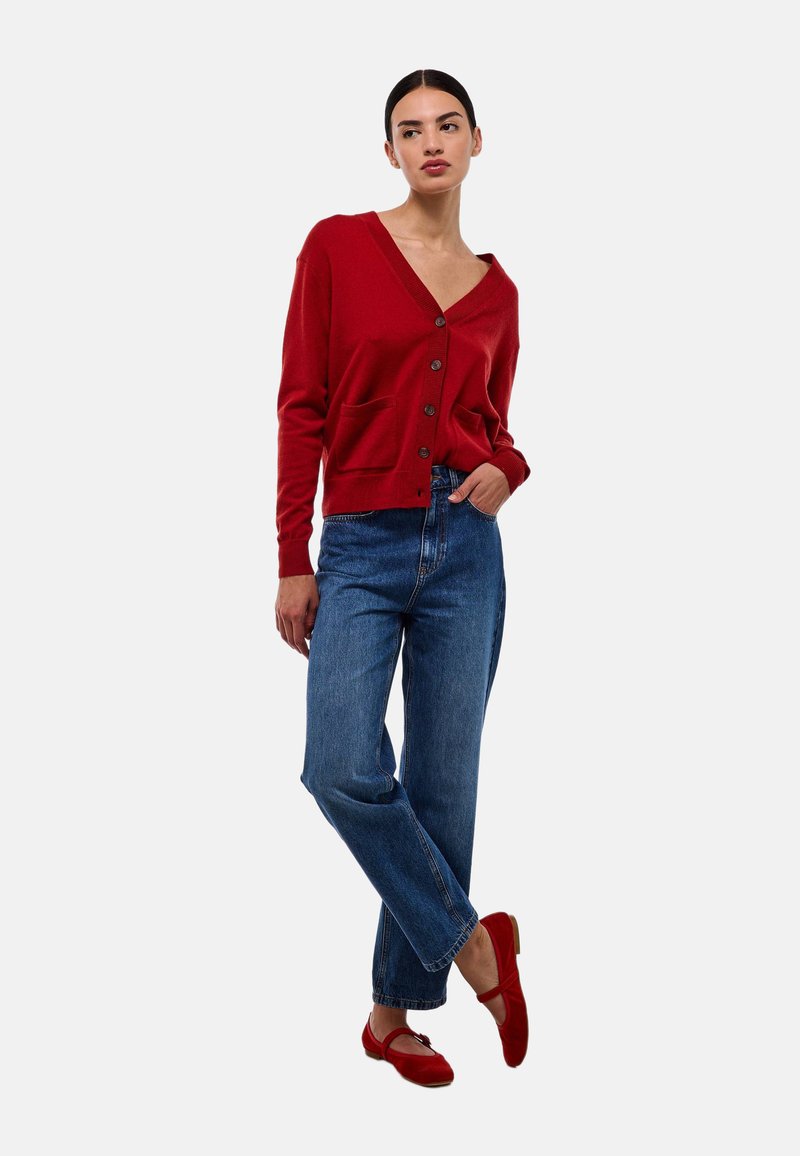 Cardigan rouge avec col en V, fermeture à boutons et poches avant, associé à un jean taille haute bleu et des ballerines rouges. Texture lisse.