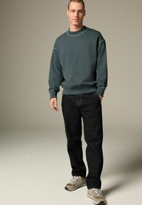 Tmavozelená mikina s rebrovanými manžetami a výstrihom, zkombinovaná s tmavými džínsami a šedými teniskami. Jednoduchý dizajn, voľný strih, minimálne detaily.