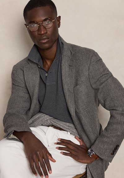 Homme portant des lunettes rondes, un polo gris, un blazer en chevron, un pantalon blanc et une montre-bracelet, assis avec une main sur ses genoux et une autre à la taille.