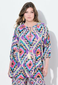 Buntfarbenes Tunika mit geometrischen Mustern in Lila, Pink und Gelb. Verfügt über einen Schlüsselaugen-Ausschnitt und Quasten-Details. Leichter Stoff, lockere Passform.