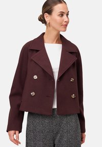 Une femme portant un manteau court bordeaux à double boutonnage avec de grands revers sur un haut blanc et un pantalon gris à motifs.