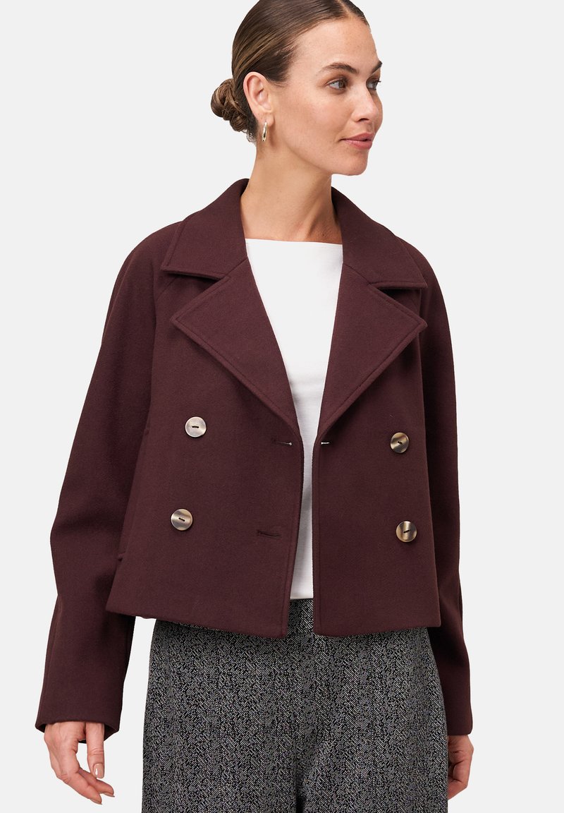 Une femme portant un manteau court bordeaux à double boutonnage avec de grands revers sur un haut blanc et un pantalon gris à motifs.