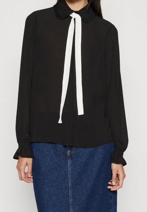 Czarna bluzka z długim rękawem z falbankowym kołnierzem i mankietami, ozdobiona białą kokardą, noszona z niebieską spódnicą dżinsową.