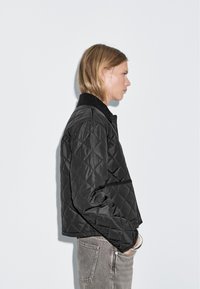 Black quilted jacket with a collar, featuring a cropped design and textured surface. Worn with gray high-waisted jeans.
