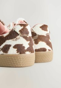 Ein Paar Sneakers mit einem Kuhfellmuster in Braun und Weiß sowie strukturierten, beige-farbenen Gummisohlen vor einem hellen, neutralen Hintergrund.