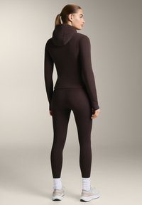 Tajmad mörkbrun hoodie med långa ärmar, med en huva. Kombinerad med matchande leggings och ljusa träningsskor.