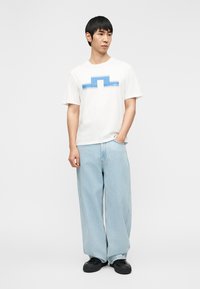 Λευκό T-shirt με κοντά μανίκια και μπλε γραφικό σχέδιο μπροστά, συνδυασμένο με ανοιχτό μπλε τζιν παντελόνι με φαρδιά μπατζάκια και μαύρα αθλητικά παπούτσια.