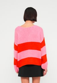 Pull en tricot présentant de larges rayures horizontales roses et rouges. Coupe carrée avec un col large et des manches amples.