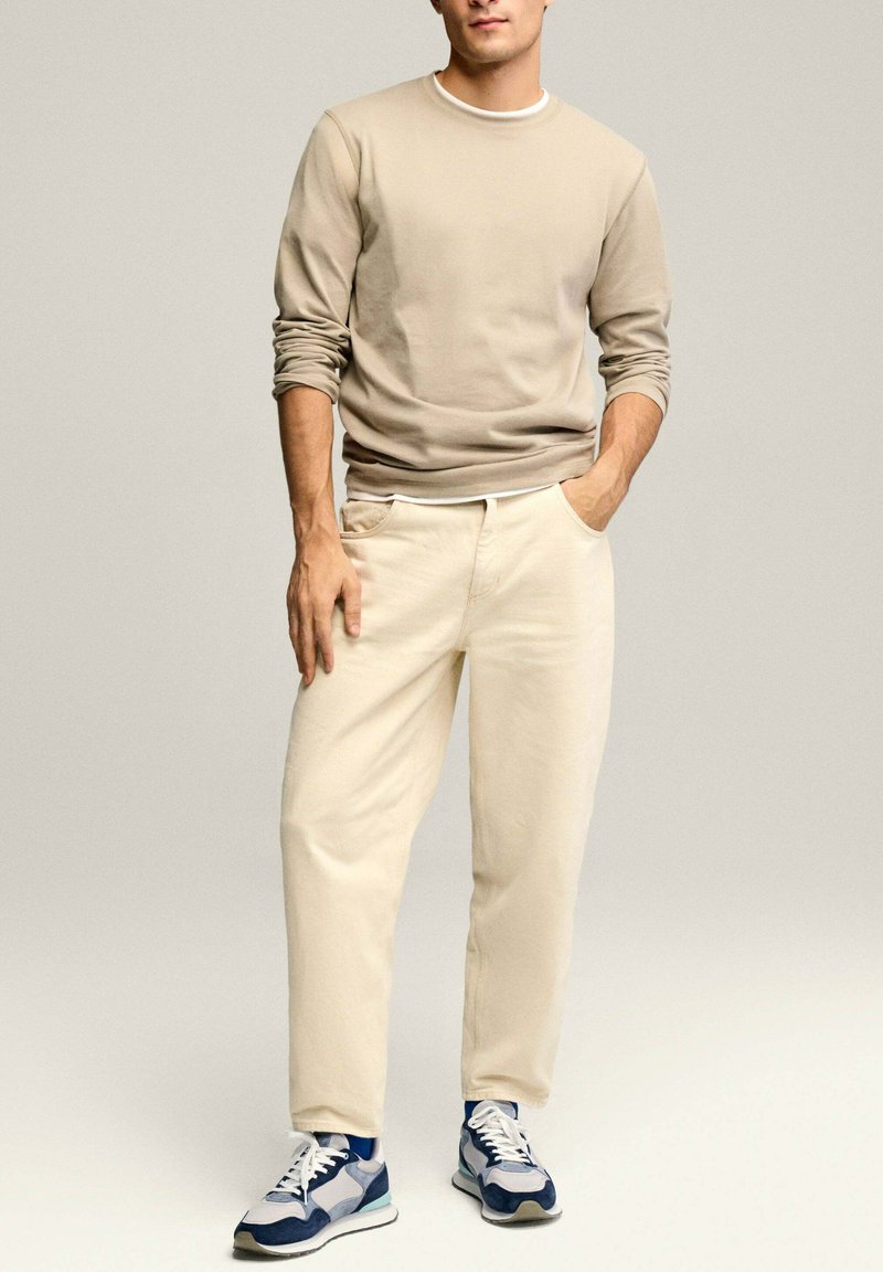 Jasno beżowa koszula z długim rękawem, luźny krój. Beżowe spodnie z kieszeniami. Niebieskie i szare sneakersy z białą podeszwą. Gładka tekstura, codzienny styl.