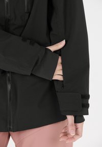 Schwarze Jacke mit einem Reißverschluss, strukturiertem Stoff, engen Bündchen mit Klettverschluss-Akzenten und einem versteckten Taschen-Detail am Ärmel.