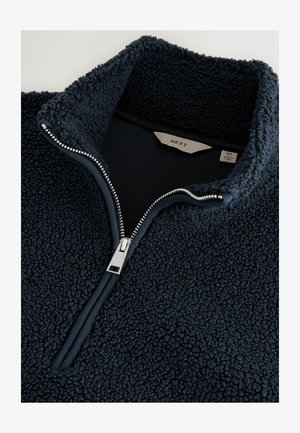 Tummansininen fleece-pullove, jossa on vetoketjukaulus, ja joka on valmistettu teksturoidusta, pehmeästä materiaalista. Vetoketjessä on hopeinen yksityiskohta. Tagi näyttää koon medium.