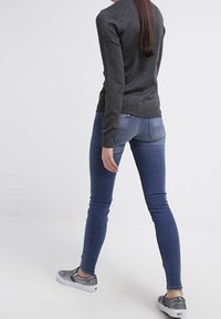 Kvinna med långt hår som bär en mörkgrå tröja, blå slim fit jeans och metalliska slip-on sneakers som går mot en enkel vit bakgrund.