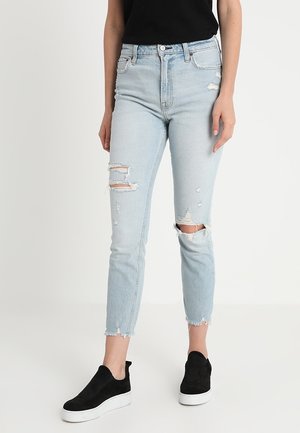 Slim fit light blue ripped jeans with frayed hems worn with black slip-on sneakers and a black top.