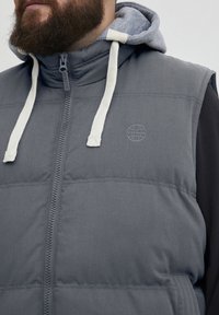 Grå polstret vest med hette og kontrasterende innerfôr, med frontlås, hvite snorer og et preget globus-logo.
