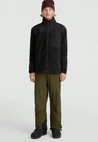 Ungdom i en svart zip-up jakke, olivengrønne bukser, svarte sko og en burgunder hue, stående mot en ensfarget lys bakgrunn.