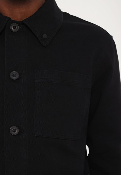 Black denim jacket featuring a button-up front, pointed collar, single chest pocket, and subtle texture, showcasing a sleek design.