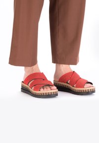 Des pieds portant un pantalon court marron et des sandales à plateforme rouges à enfiler avec des semelles en jute rayées sur un fond blanc.