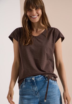 Femme souriante, portant un haut rayé marron à manches courtes avec un nœud sur le côté et un jean bleu, debout devant un fond neutre.