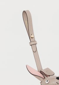 Šviesiai béžinė odinė juosta su lygia tekstūra. Yra rožinė ausis ir maža juoda akytė, užsegta auksinės spalvos snapeliu.