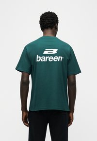 Άνδρας με πλάτη προς το φακό, φοράει σκούρο πράσινο T-shirt με μεγάλο λευκό λογότυπο «B bareen» στην πλάτη και μαύρο παντελόνι, μπροστά σε λευκό φόντο.