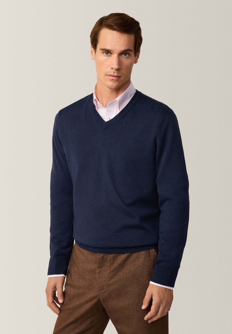 Maglione blu navy a scollo a V in morbido tessuto, abbinato a una camicia color rosa chiaro con colletto. Presenta polsini a costine e una texture liscia.
