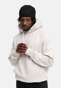 Hellbeige Kapuzenpulli mit einer Kängurutasche, Zugbändern und gerippten Bündchen, getragen mit einer schwarzen Mütze. Frontalansicht, Hände gefaltet.