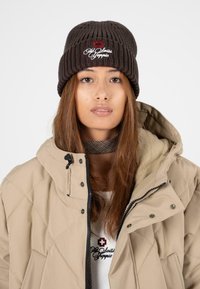 Bruine geribbelde gebreide beanie met geborduurd logo, gecombineerd met een beige gewatteerde jas. Heeft een hoge kraag en contrasterende zwarte rits.