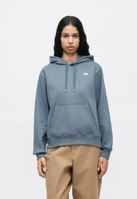 Γυναίκα με μακριά σκούρα μαλλιά, φορώντας μπλε hoodie της The North Face και μπεζ παντελόνι, που στέκεται μπροστά σε λευκό φόντο.