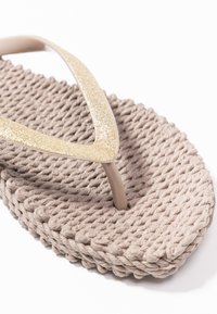 Beige flip-flop with a textured woven sole and glossy gold glitter straps, featuring a V-shaped design and soft, flexible material.