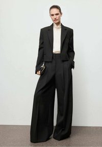 Zwarte getailleerde blazer met schoudervullingen, gecombineerd met high-waisted wijde pantalons. Een witte blouse en een kleine zwarte clutch maken de look compleet.