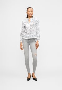 Γυναίκα που στέκεται φορώντας λευκό πουκάμισο με μακριά μανίκια και λουλούδια, γκρι ανοιχτό σκισμένο στενό τζιν και μαύρες μυτερές γόβες σε λευκό φόντο.