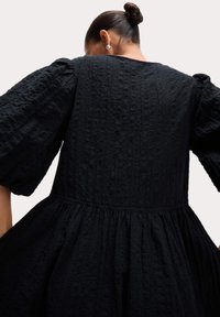 Schwarzes Kleid mit texturiertem, gecrinkeltem Stoff, gepufften langen Ärmeln und einer gerafften Taille, die eine fließende Silhouette schafft.