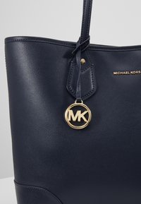 Sac cabas en cuir texturé bleu marine avec logo Michael Kors doré et pendentif circulaire MK suspendu à la poignée.
